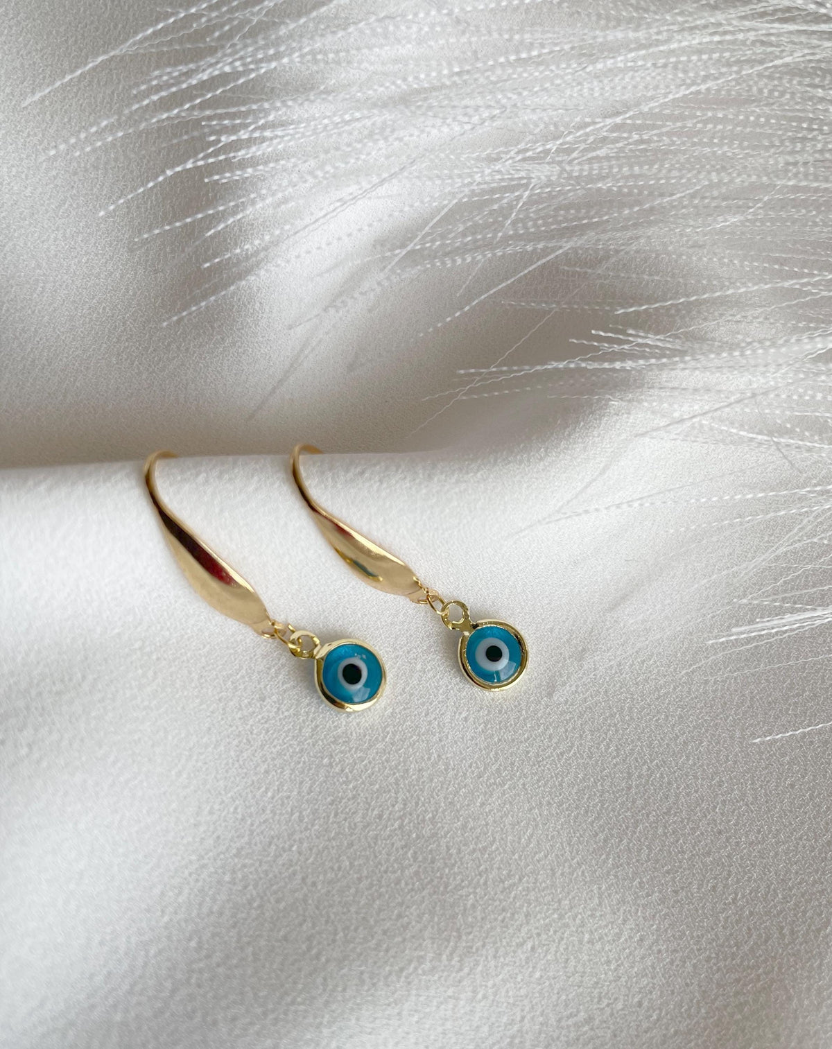 A pair of gold plated drop earrings with circular blue and black evil eye charms, displayed on a textured white fabric background.