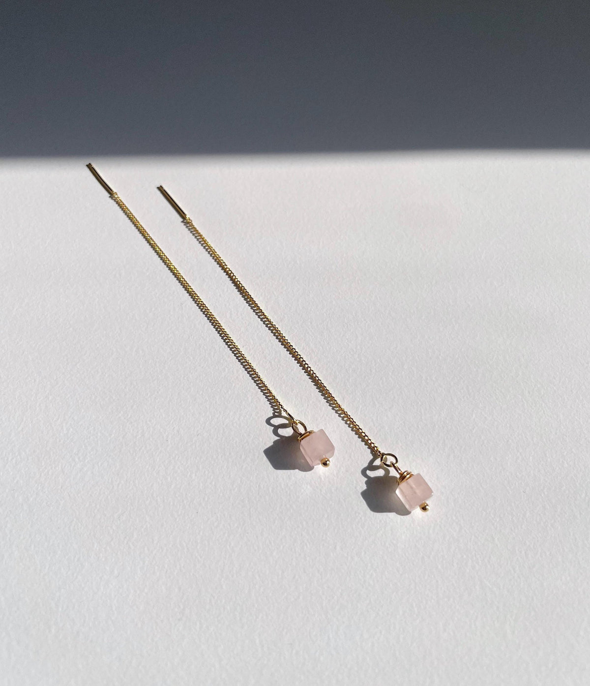 A pair of long gold chain threader earrings with small pale pink rose quartz square bead charms, photographed on a white surface with a dark shadow across the upper background.