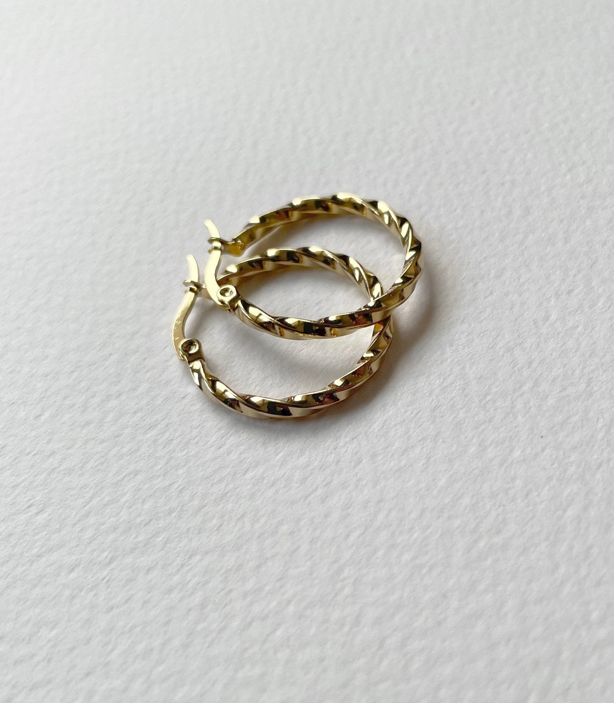 A pair of small gold hoop earrings with a textured, twisted surface, resting on a textured white paper background.