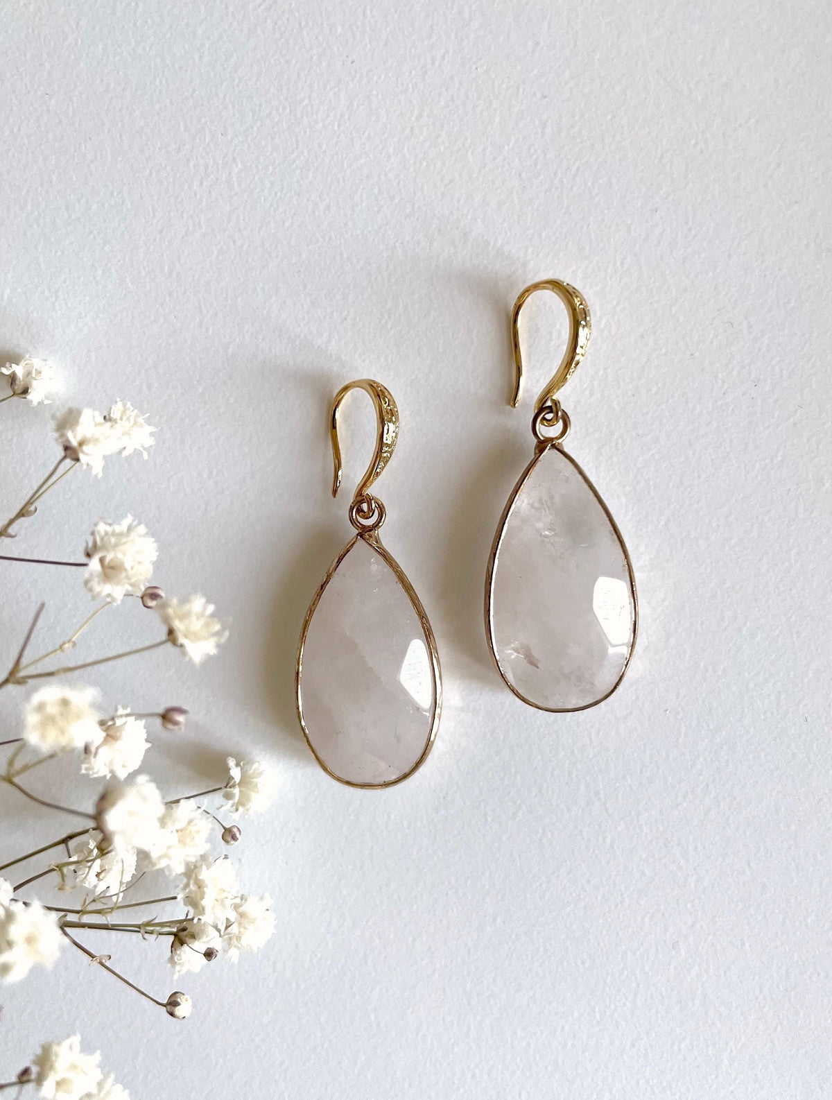 A pair of gold drop earrings with teardrop-shaped clear faceted stones, displayed on a light textured background next to small white flowers.