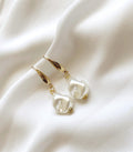 A pair of gold drop earrings with irregular white pearl-like beads, displayed on a draped white fabric background.