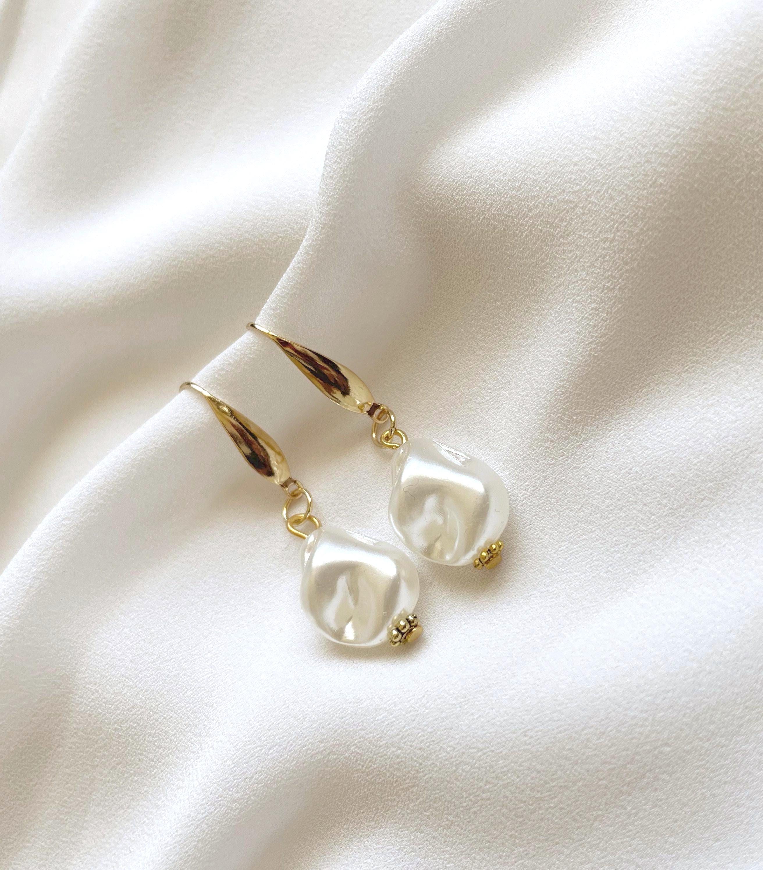 A pair of gold drop earrings with irregular white pearl-like beads, displayed on a draped white fabric background.