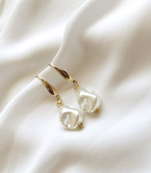 A pair of gold drop earrings with irregular white pearl-like beads, displayed on a draped white fabric background.