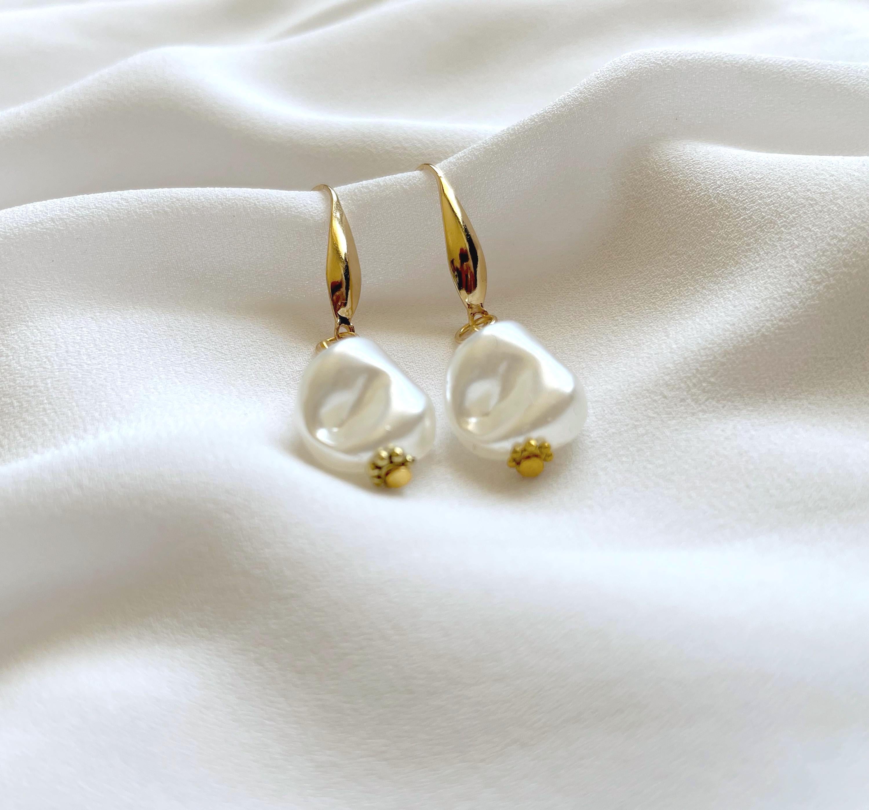 A pair of gold drop earrings with irregular white pearl dangles and small gold bead accents, displayed on draped white fabric.