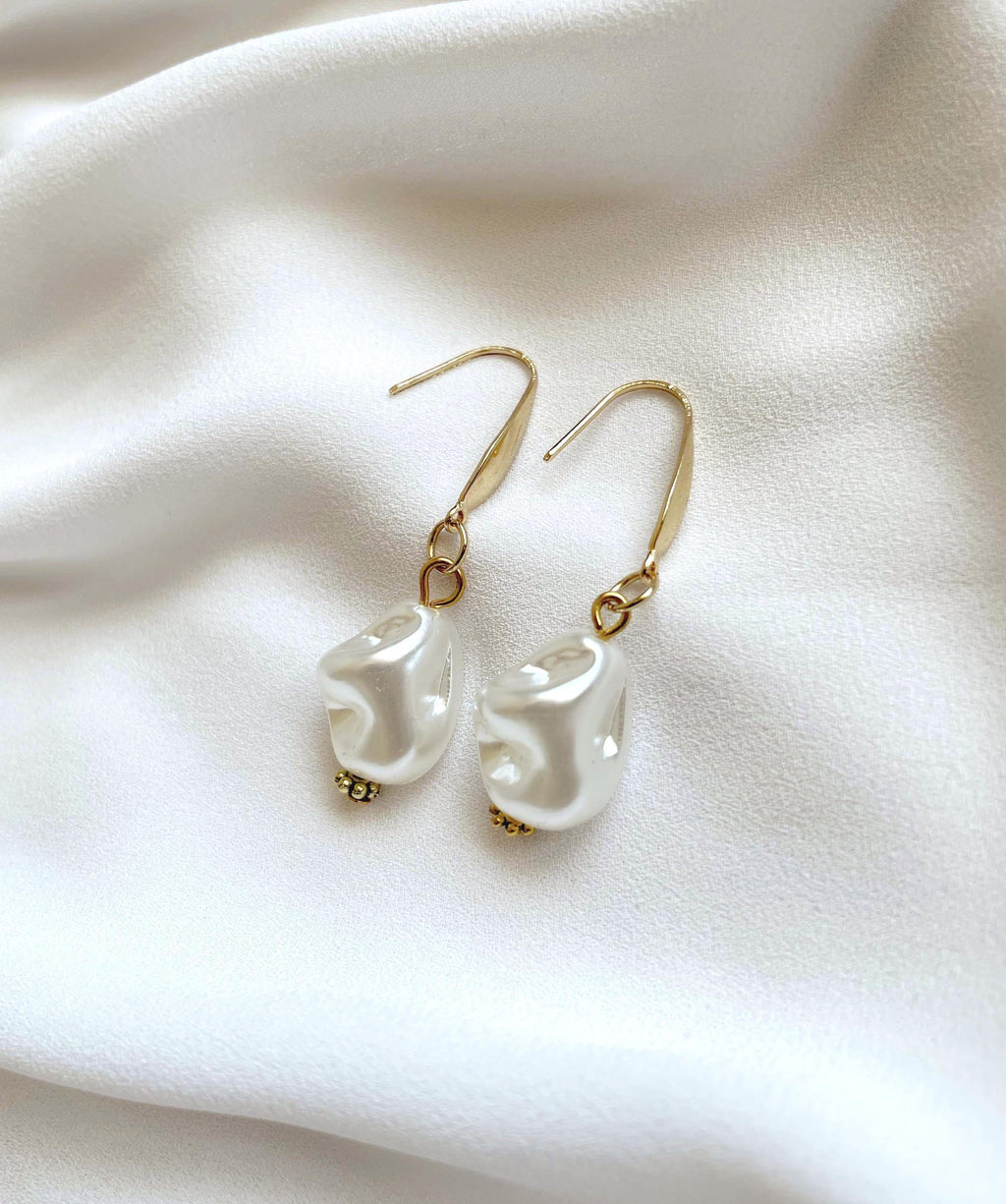 A pair of gold-tone dangle earrings with irregular white pearl drops, displayed on a soft white fabric background.