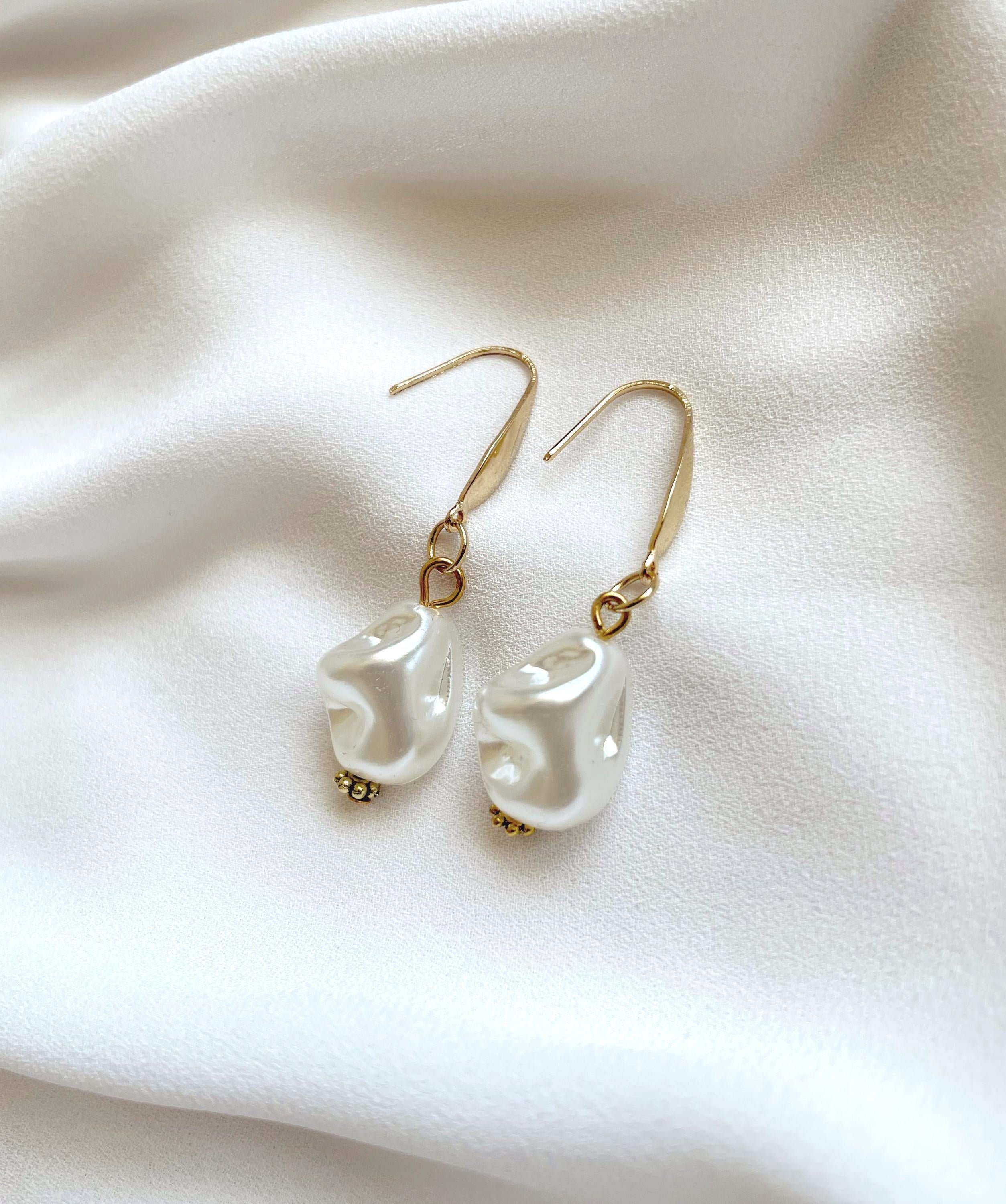 A pair of gold-tone dangle earrings with irregular white pearl drops, displayed on a soft white fabric background.