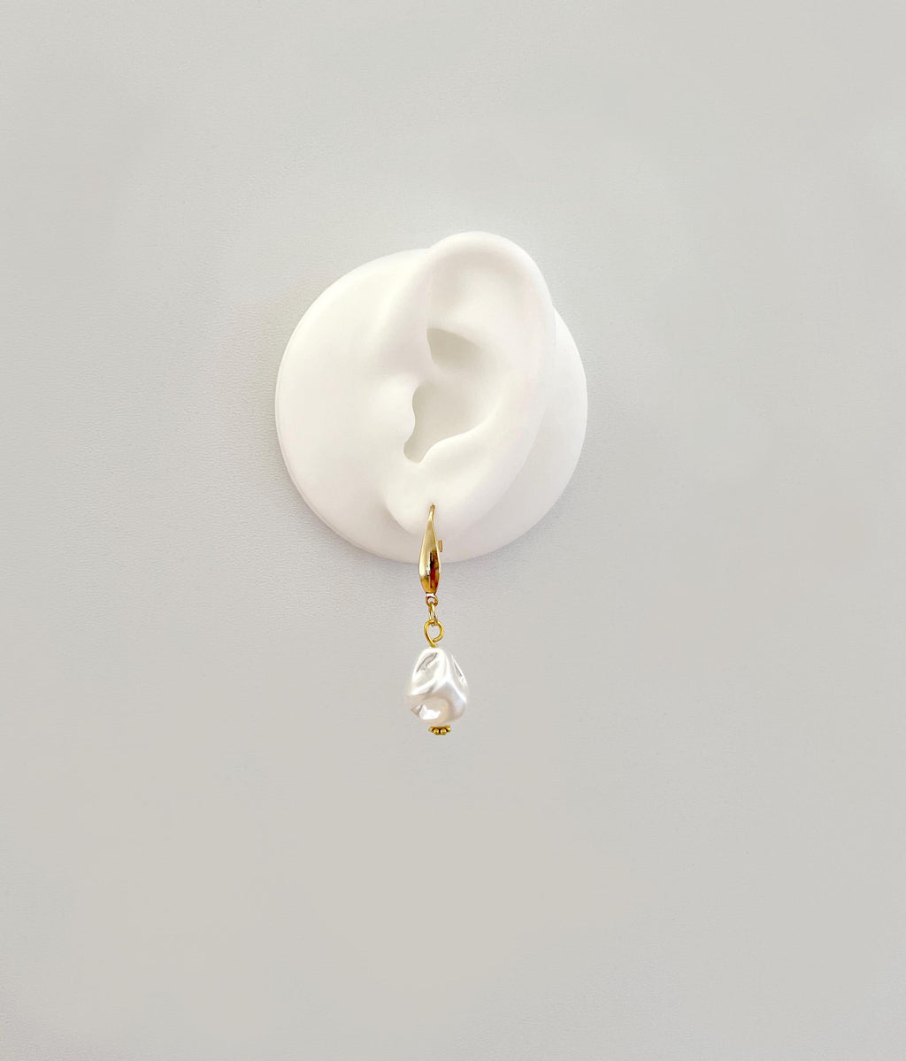A single gold hoop earring with a dangling clear teardrop-shaped pearl, displayed on a white silicone ear stand against a light gray background.