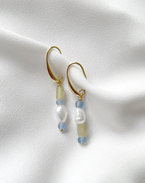 A pair of gold plated dangle earrings with baroque pearl beads, light blue beads, and pale yellow-green beads, displayed on a textured white fabric background.