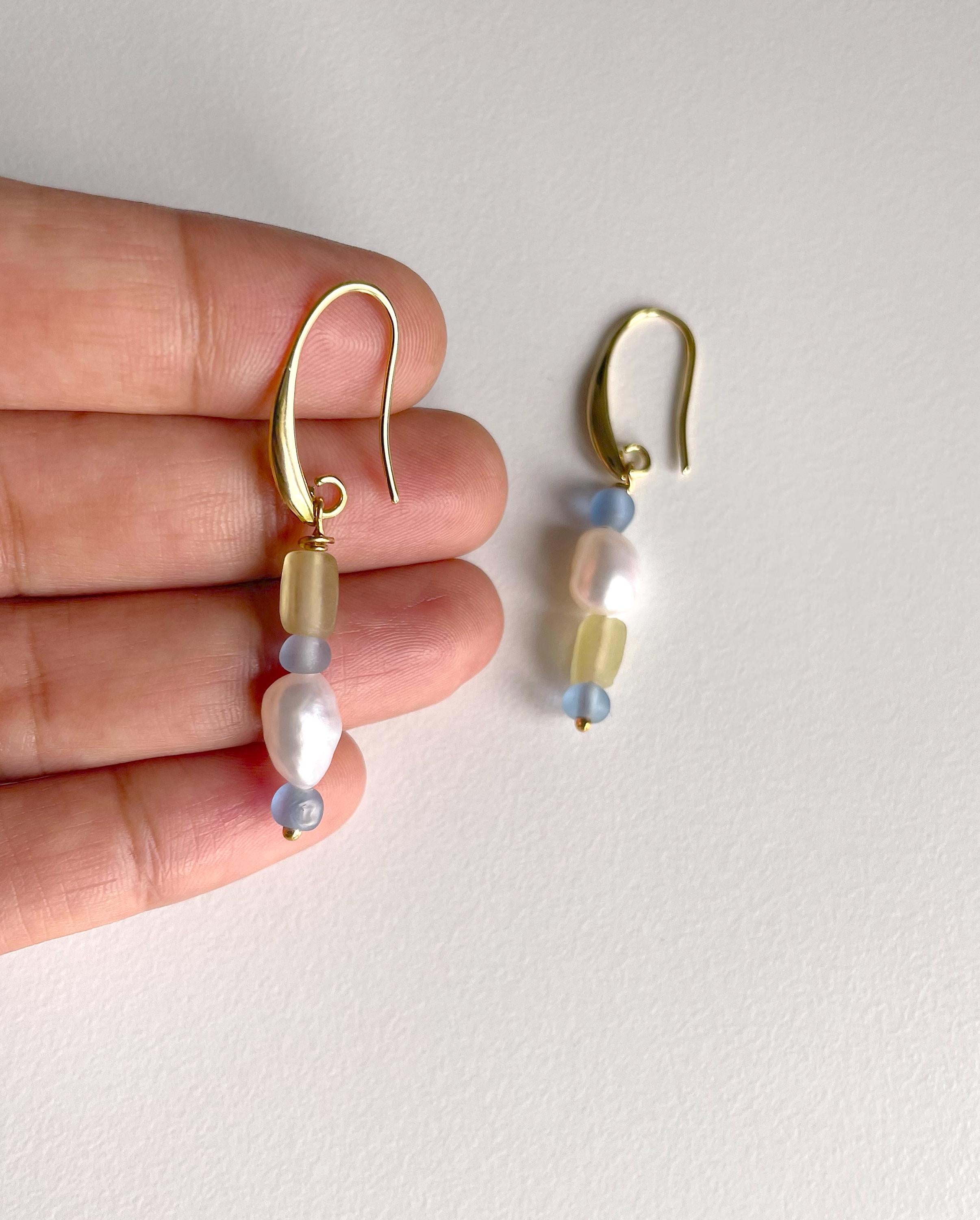 A pair of gold dangle earrings with white baroque pearls, pale yellow beads, and light blue beads, shown with a person's hand holding one earring against a plain off-white background.