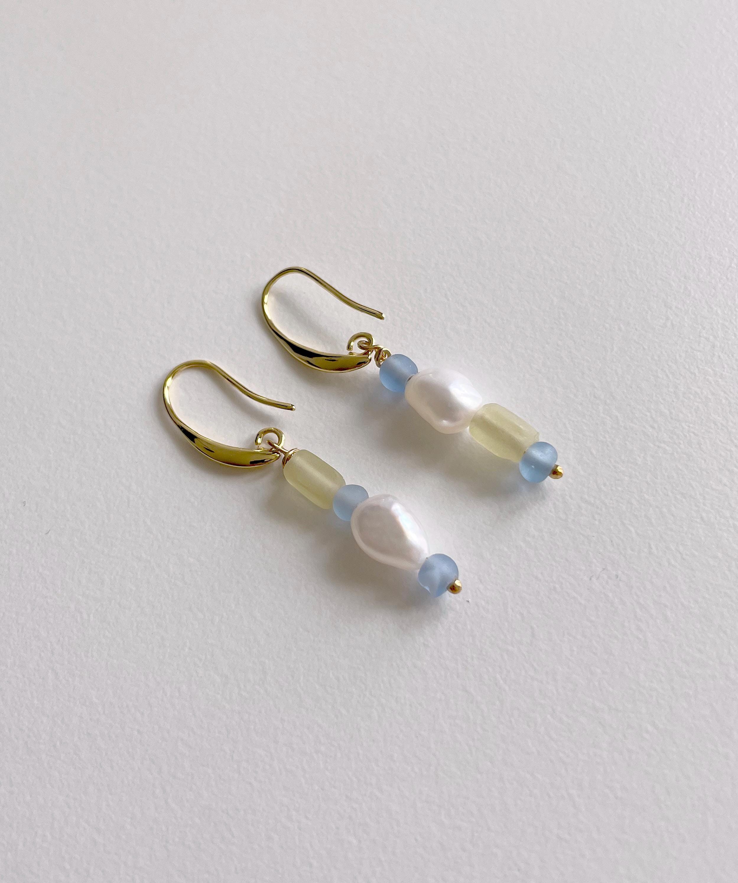 A pair of gold-tone dangle earrings with a curved hook design, featuring translucent pale yellow beads, light blue round beads, and a central white baroque pearl, displayed on a textured off-white surface.