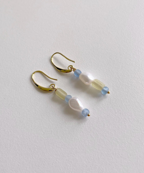 A pair of gold-tone dangle earrings with a curved hook design, featuring translucent pale yellow beads, light blue round beads, and a central white baroque pearl, displayed on a textured off-white surface.