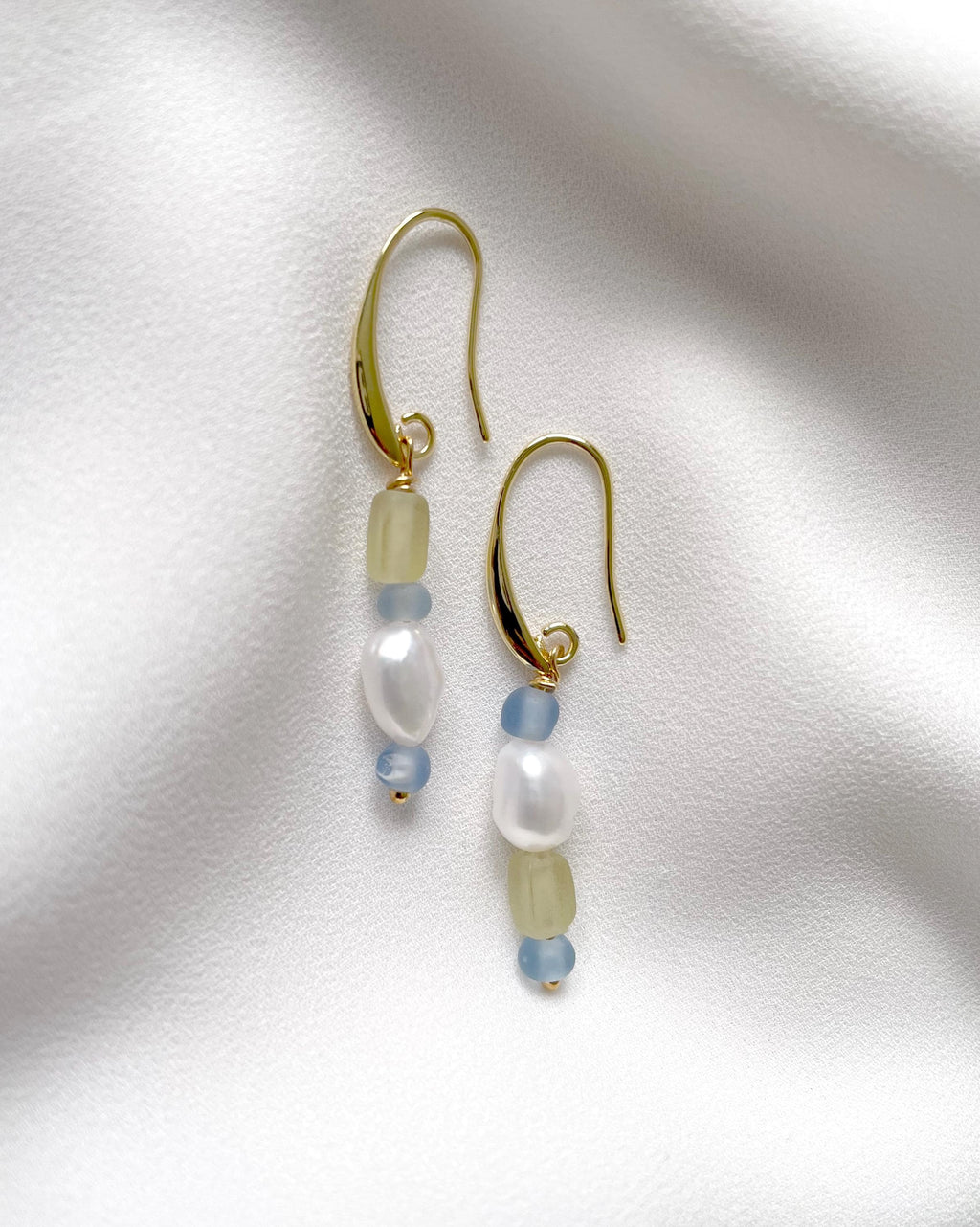 A pair of gold dangle earrings with white baroque pearls, pale green beads, and light blue beads, displayed on a textured white fabric background.