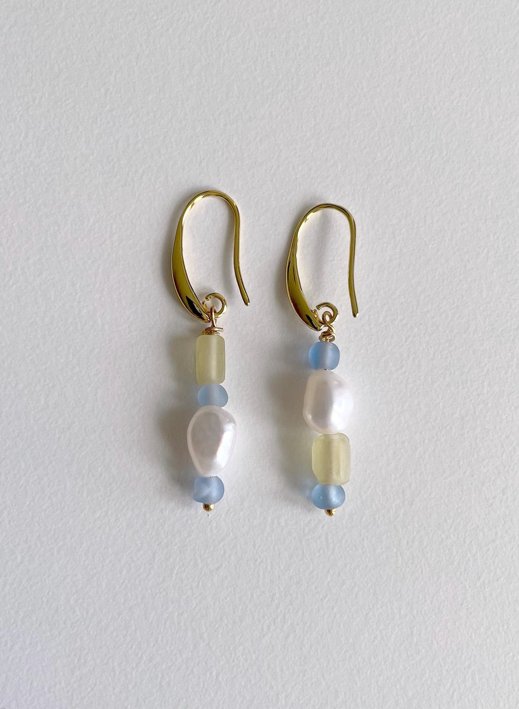 A pair of gold-tone dangle earrings with pale blue and cream beads, displayed on a textured off-white background.
