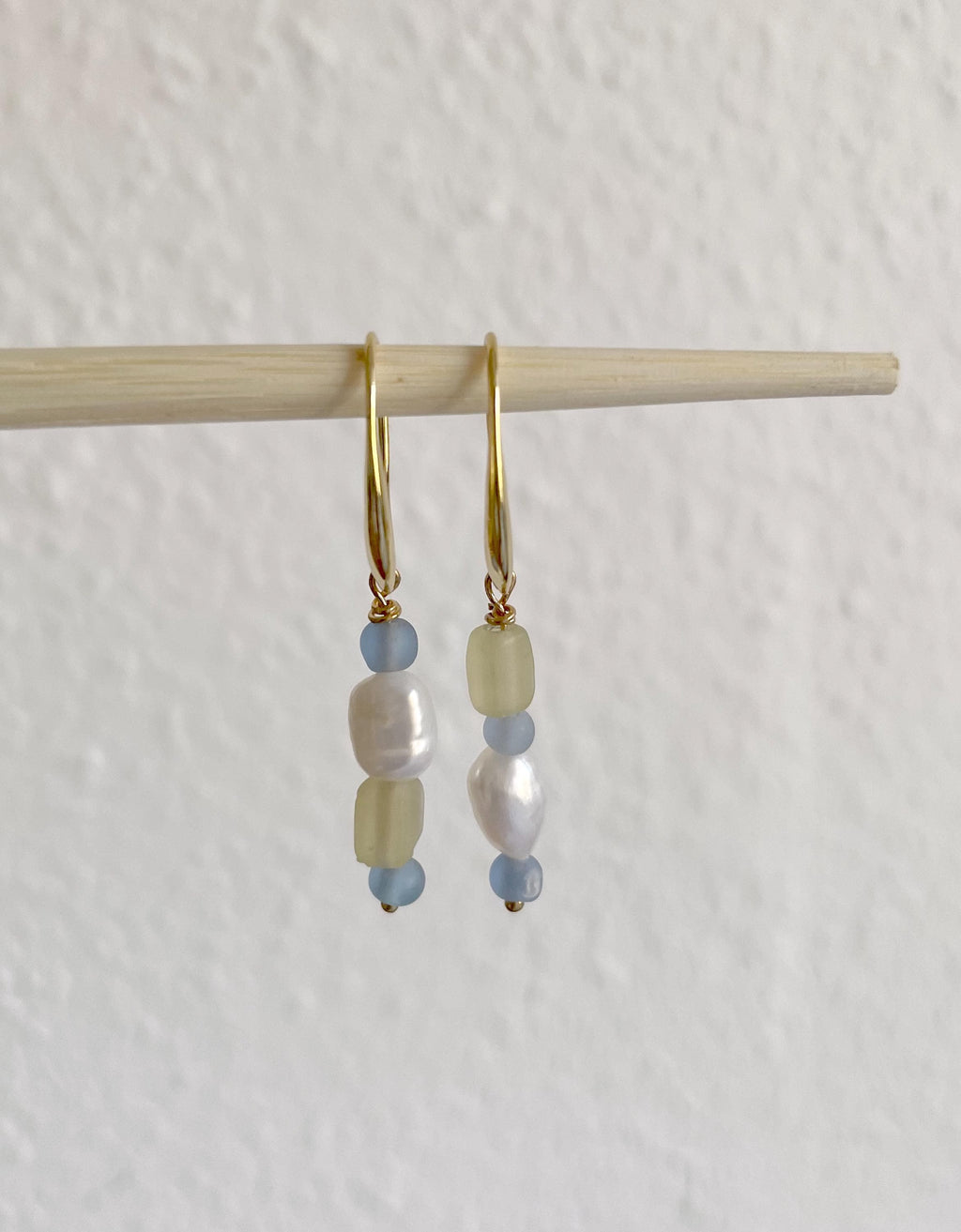 A pair of gold hoop earrings with baroque pearls, light blue beads, and pale yellow beads, hanging from a wooden stick against a textured off-white wall.