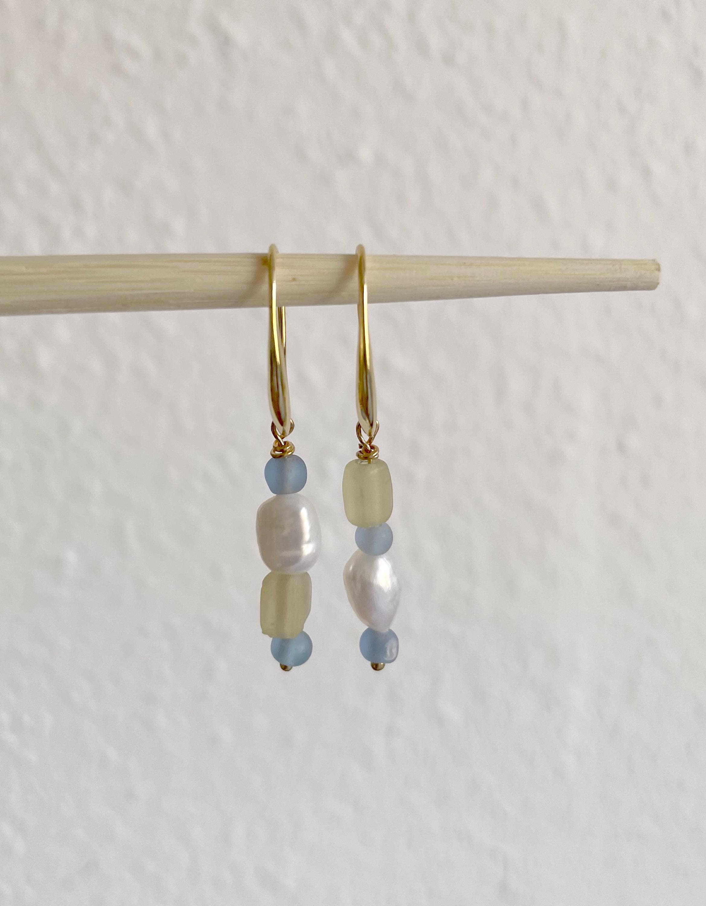 A pair of gold hoop earrings with baroque pearls, light blue beads, and pale yellow beads, hanging from a wooden stick against a textured off-white wall.