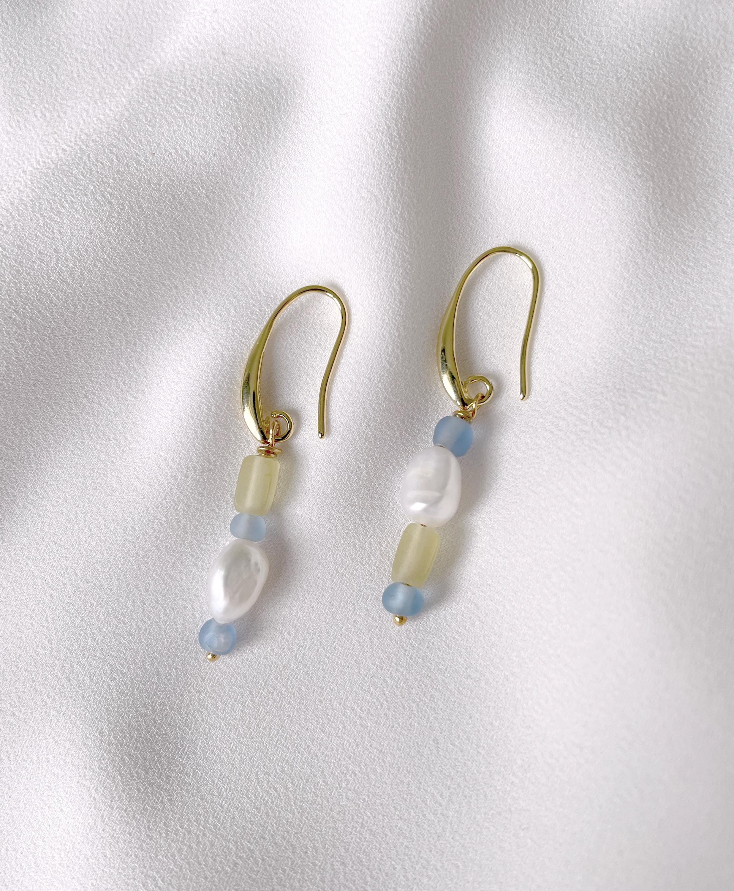 A pair of gold-tone dangle earrings with translucent pale yellow beads, light blue beads, and a white pearl, displayed on a textured white fabric background.