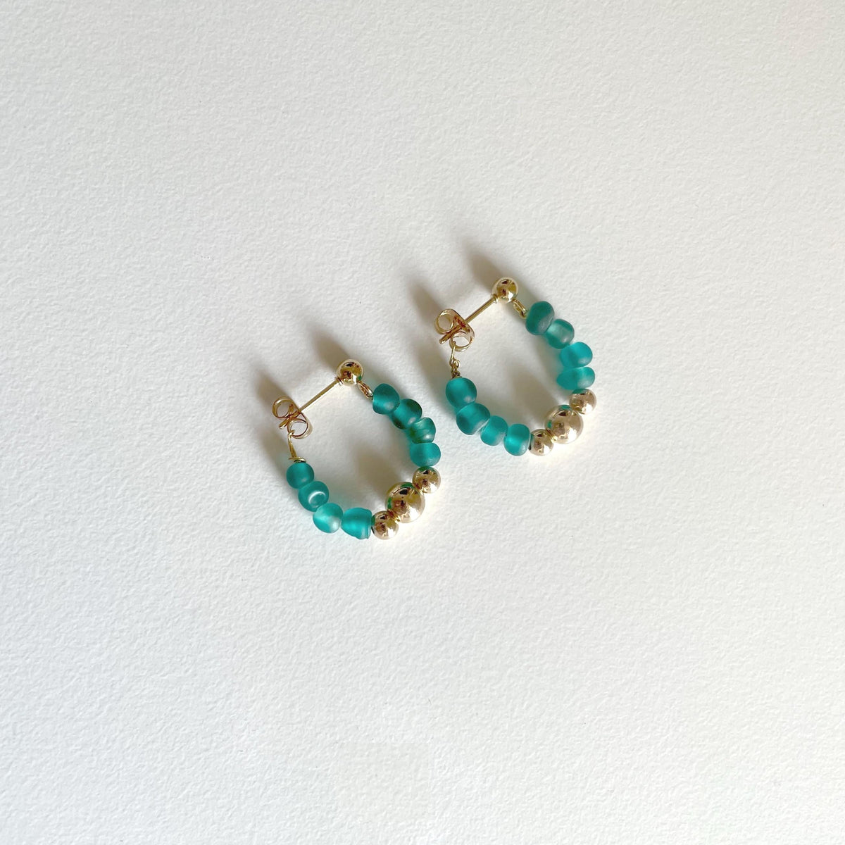 A pair of small gold hoop earrings with beaded turquoise and gold accents, displayed on a textured white surface.