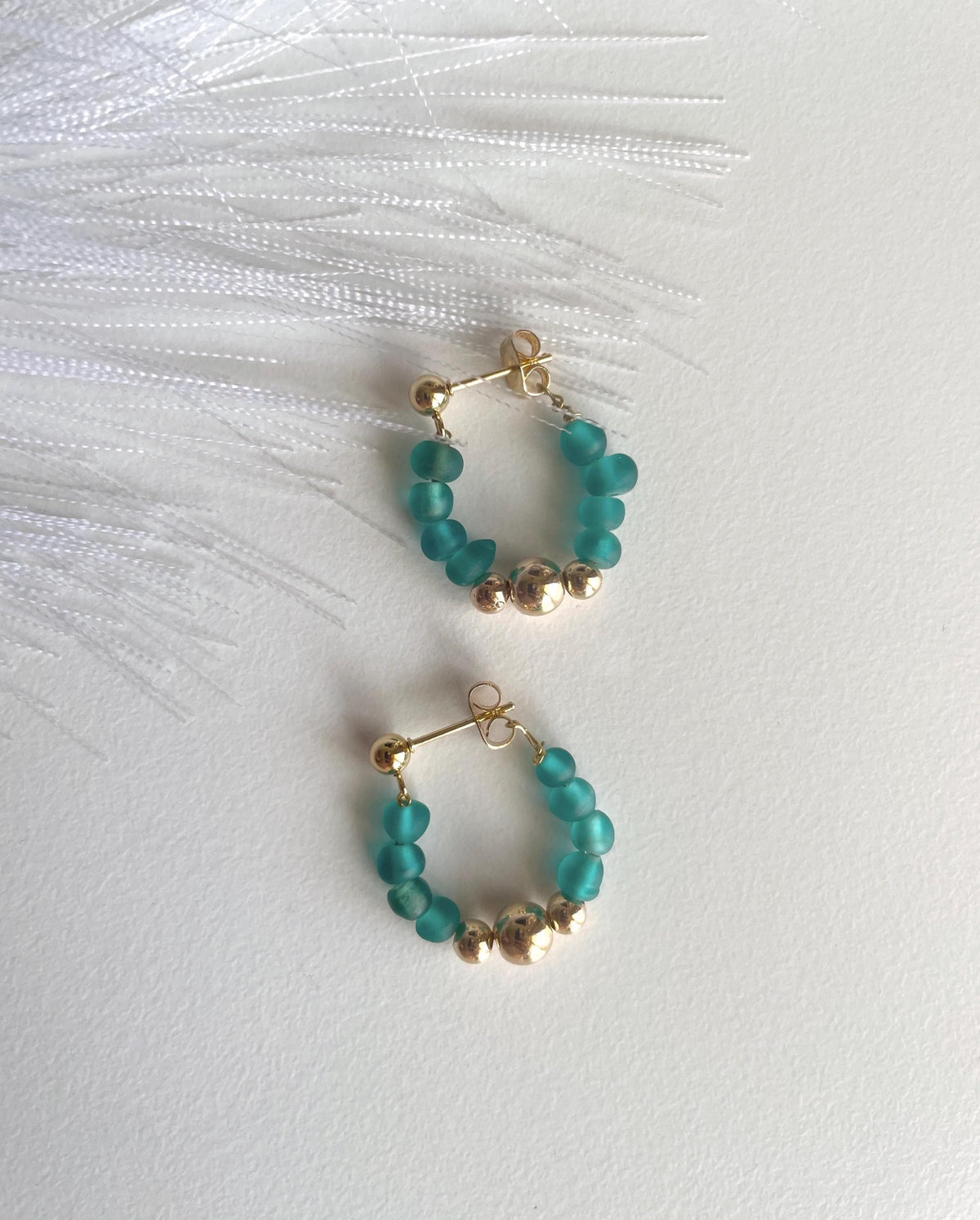 A pair of gold hoop earrings with small teal beads and metallic gold accent beads, displayed on a white textured surface with white feathers.