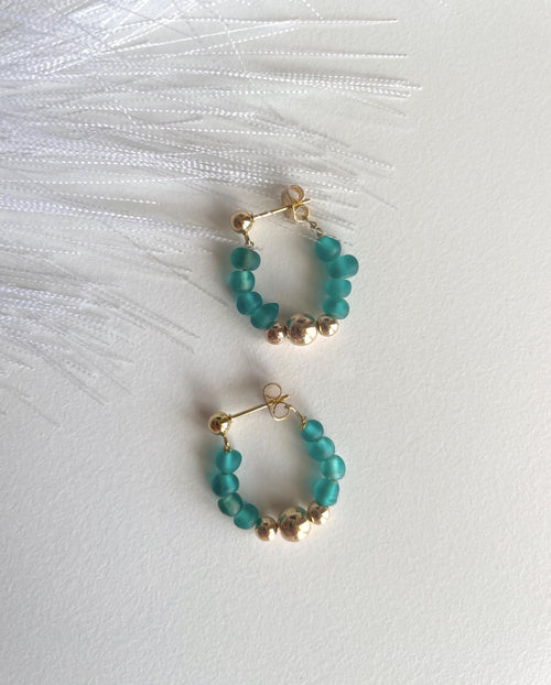 A pair of gold hoop earrings with small teal beads and metallic gold accent beads, displayed on a white textured surface with white feathers.
