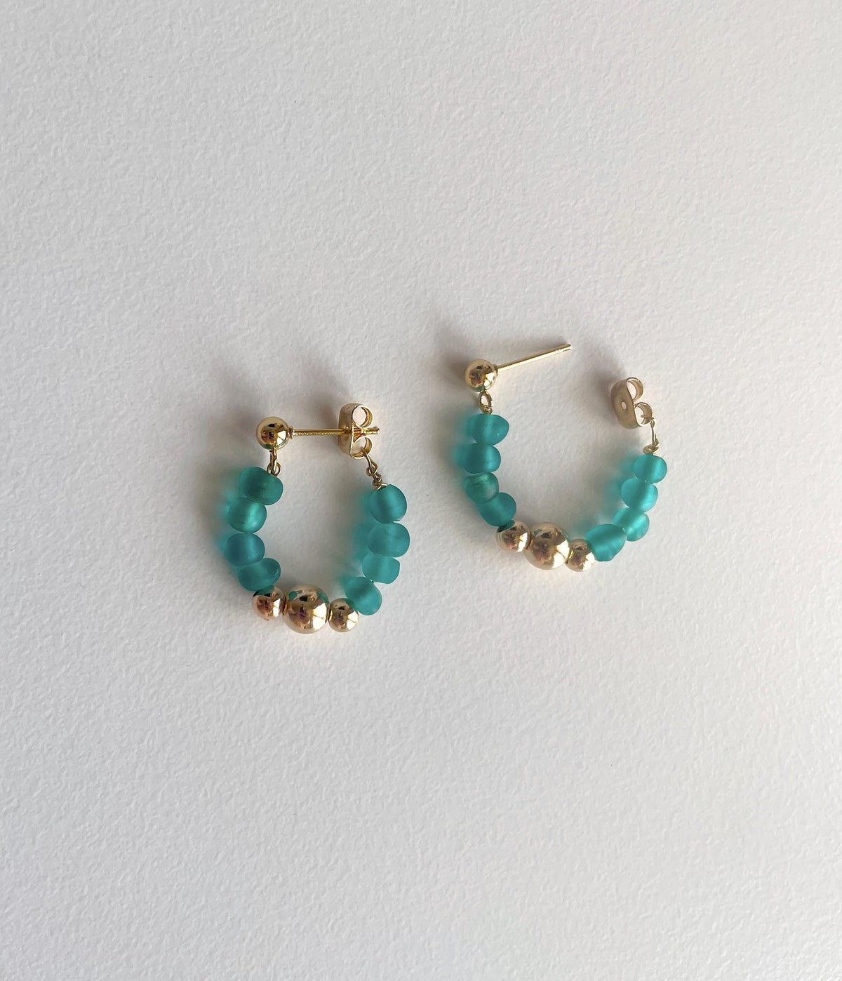 A pair of gold hoop earrings with small faceted teal beads and a few metallic gold beads, displayed on a light gray textured background.