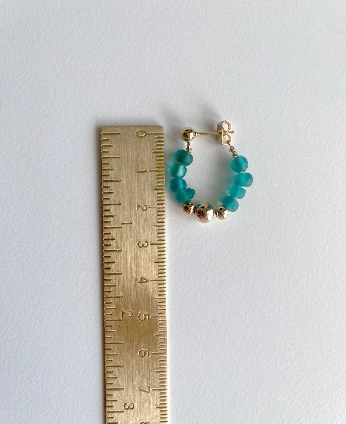 A pair of small hoop earrings with teal beads and gold-toned metal beads, shown next to a wooden ruler on a plain light background.