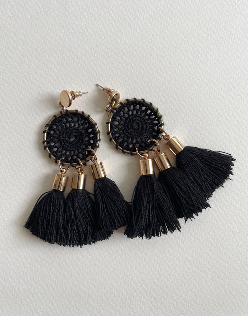 A pair of black circular crochet tassel earrings with gold-tone metal accents, displayed on a textured off-white background.