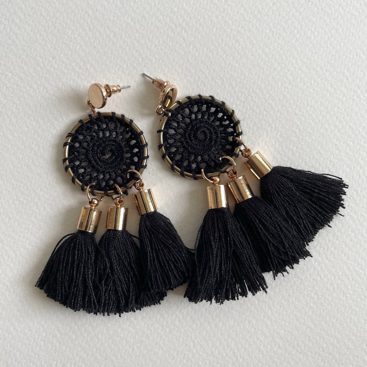 A pair of black circular crochet tassel earrings with gold-tone metal accents, displayed on a textured off-white background.