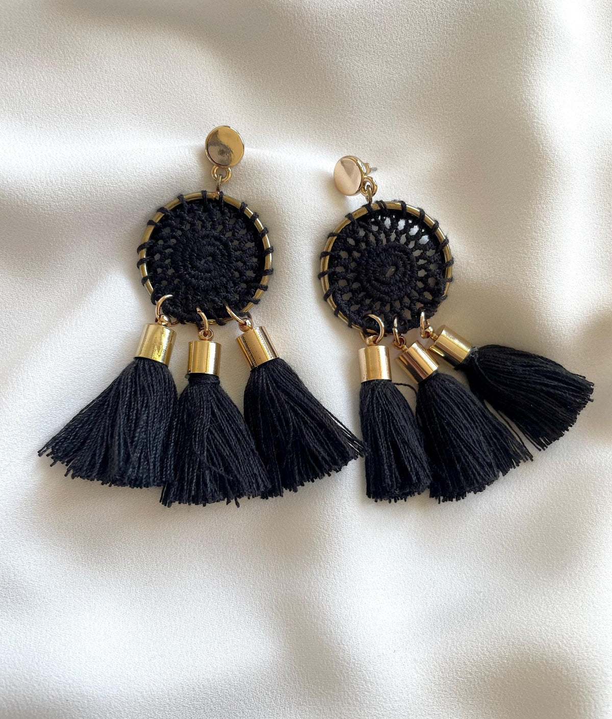 A pair of black bohemian crochet dangle earrings with gold-tone metal accents and three black tassels, displayed on a white satin fabric background.