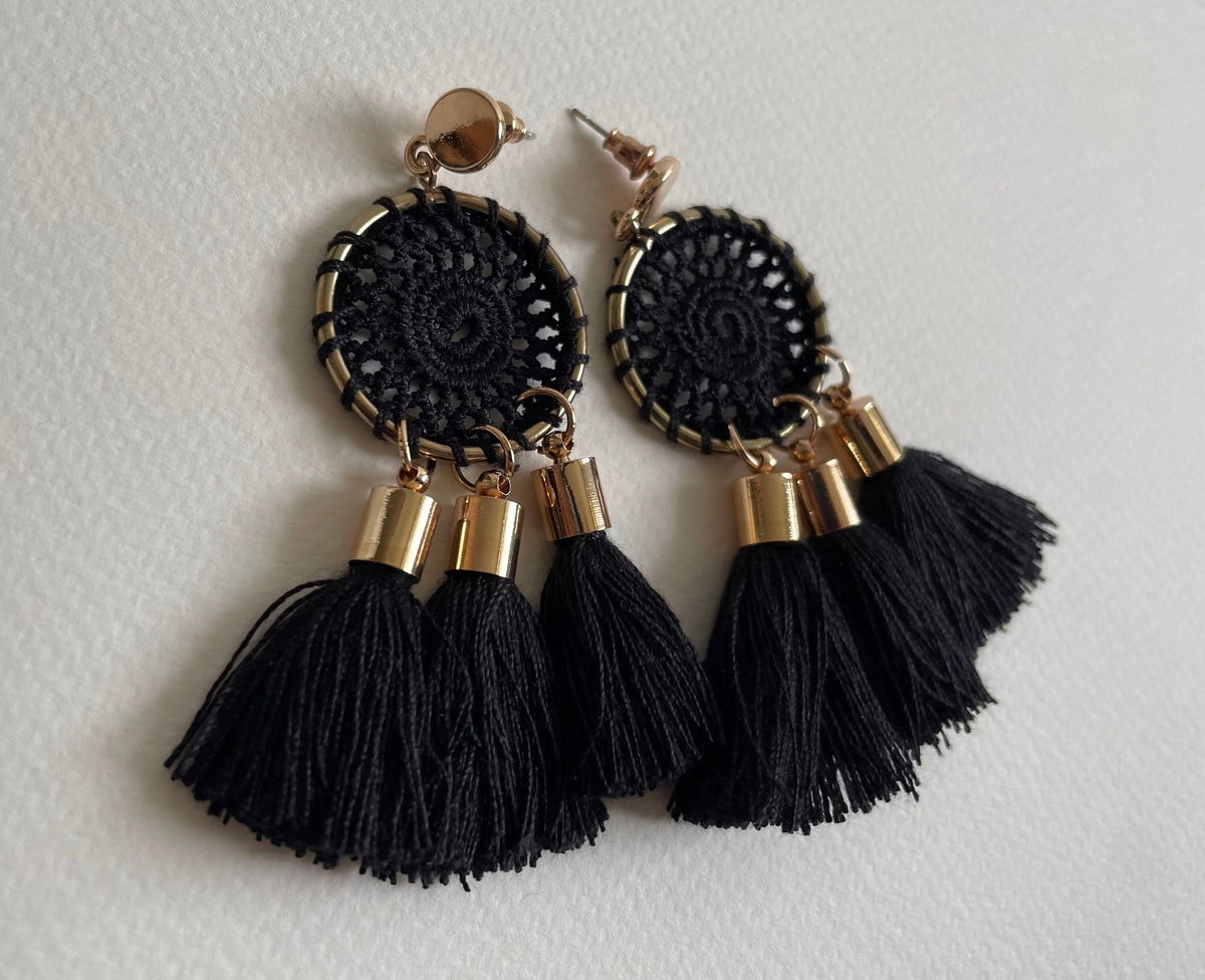 A pair of black crocheted dangle earrings with gold-tone metal accents and long black tassels, displayed on a light textured background.