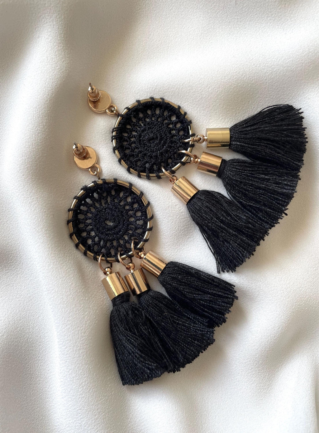 A pair of bohemian-style dangle earrings featuring black crocheted circular dreamcatcher designs, gold-tone metal accents, and three black tassels, displayed on a white satin fabric background.