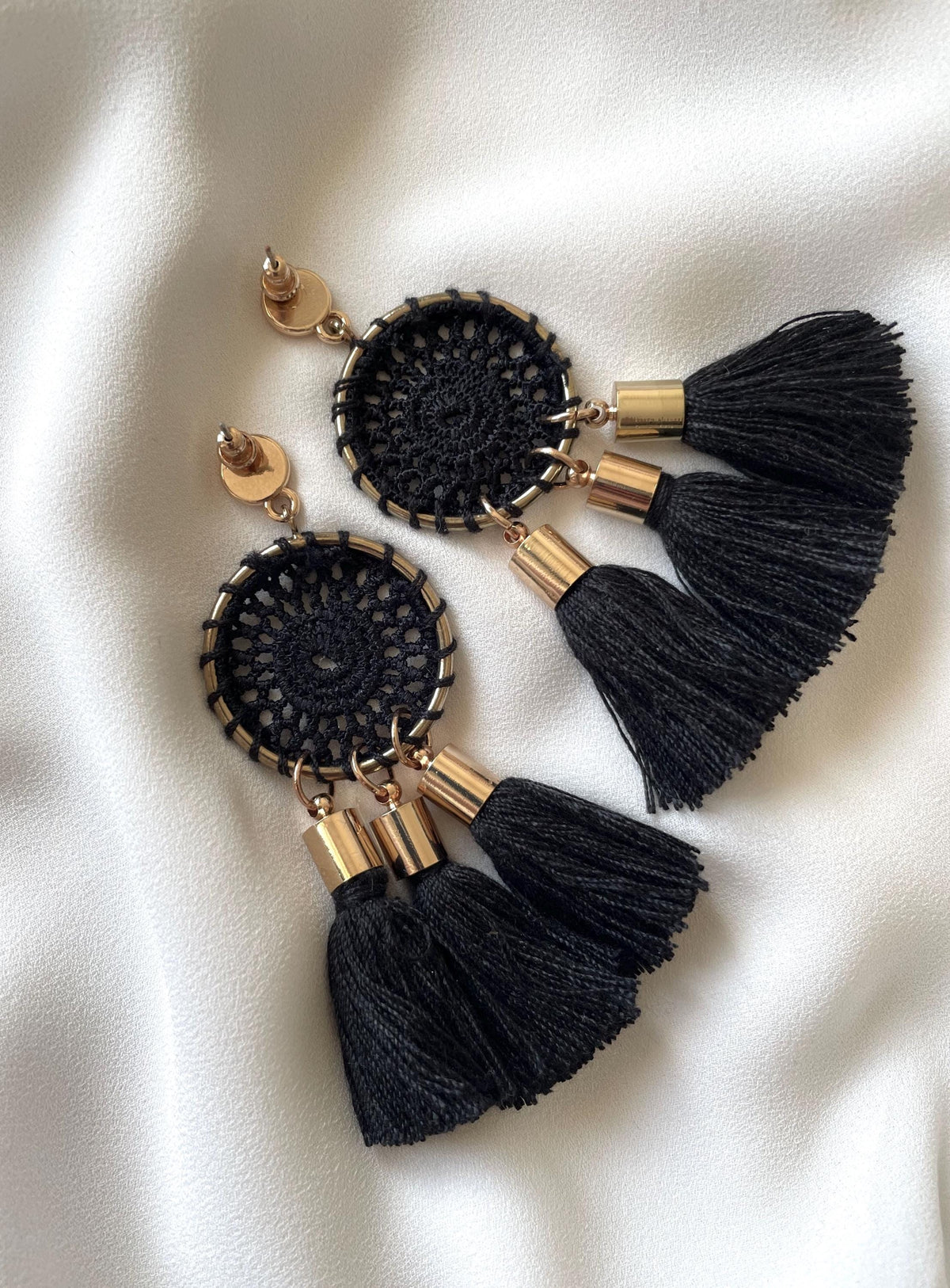 A pair of bohemian-style dangle earrings featuring black crocheted circular dreamcatcher designs, gold-tone metal accents, and three black tassels, displayed on a white satin fabric background.