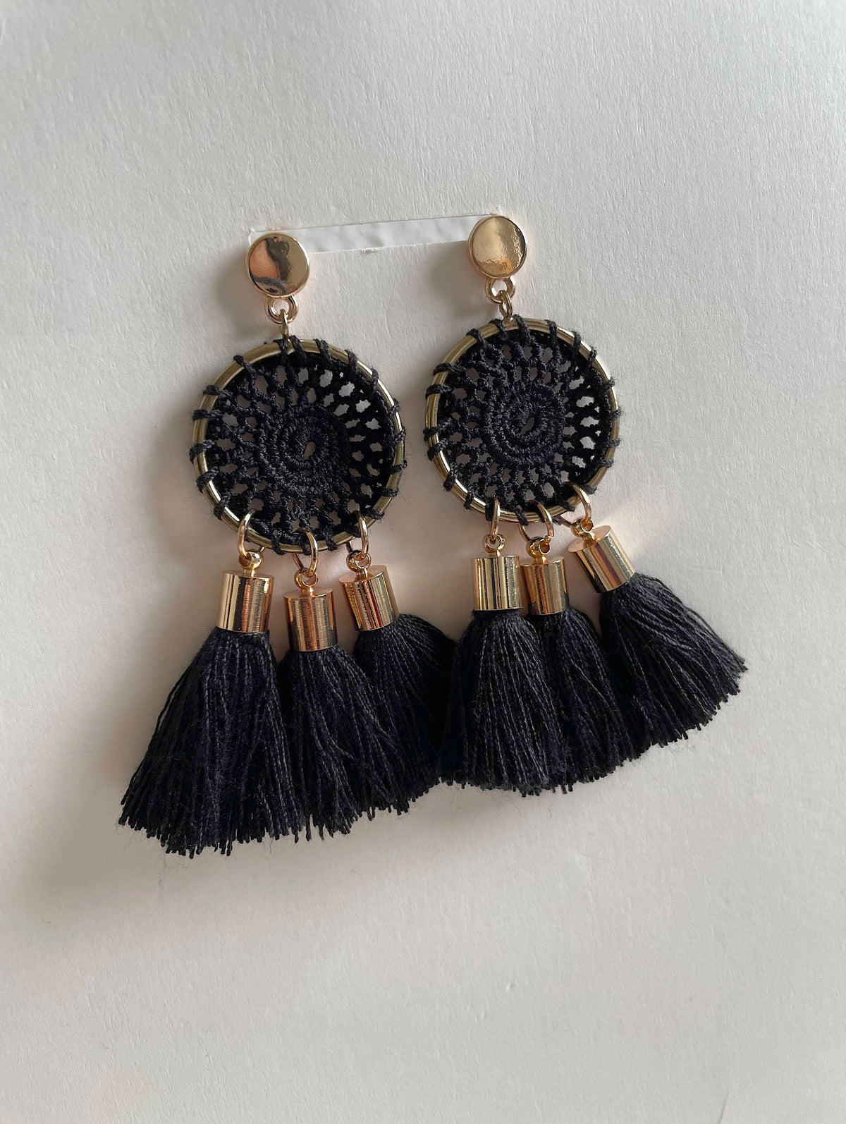 A pair of black and gold statement earrings featuring circular woven black top pieces, gold-toned metal caps, and three black tassels hanging from each earring, displayed against a plain off-white background.