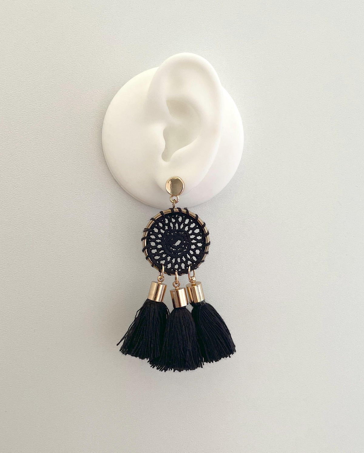 A single black and gold tassel dangle earring displayed on a white ear-shaped stand against a light gray background.