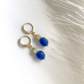A pair of gold plated hoop earrings with small rectangular sage green amazonite beads and round blue jade beads dangling below, photographed on a white surface with soft white feather-like decor.