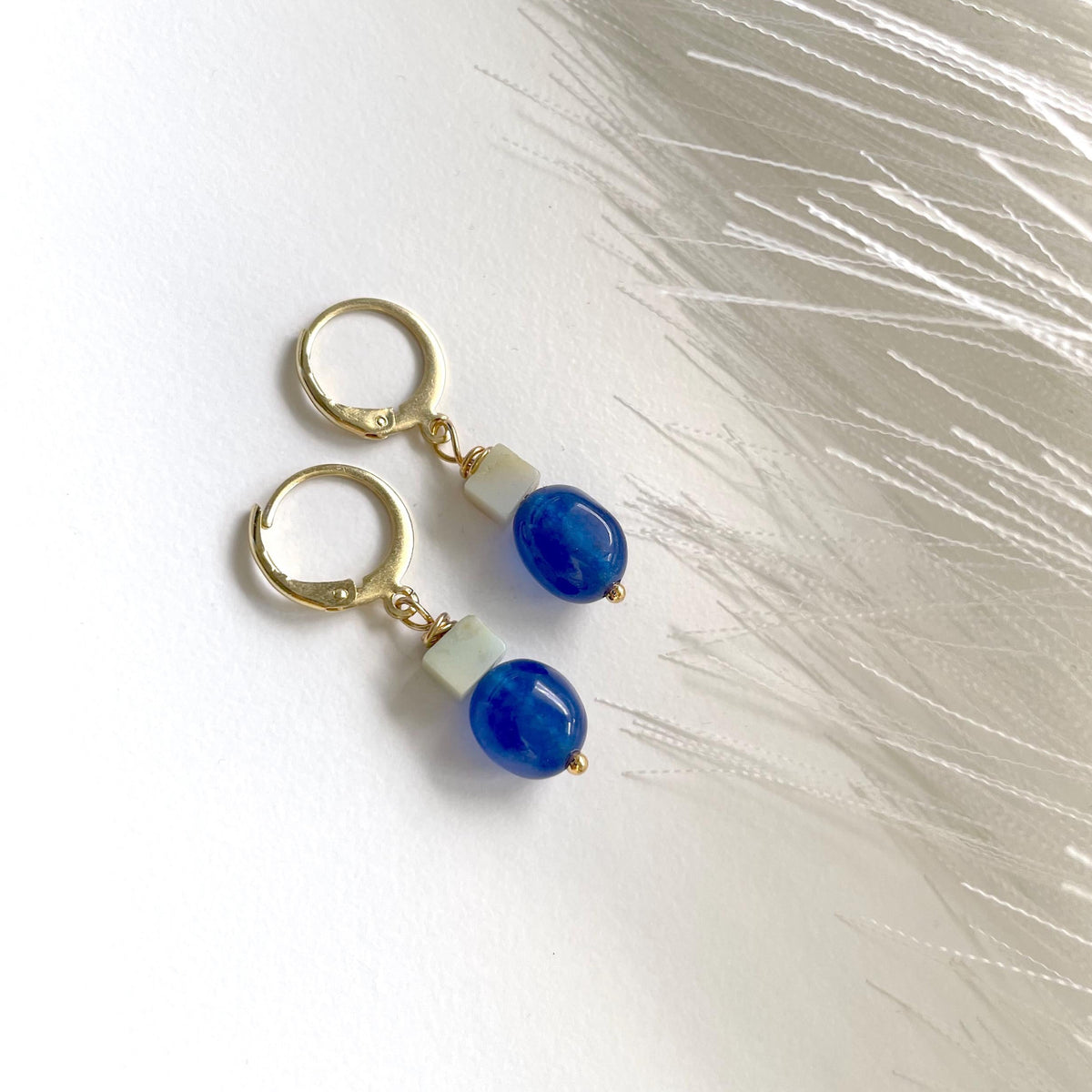 A pair of gold plated hoop earrings with small rectangular sage green amazonite beads and round blue jade beads dangling below, photographed on a white surface with soft white feather-like decor.