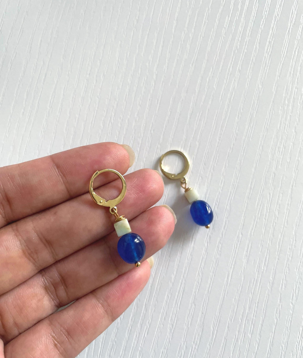 A pair of gold plated hoop earrings with blue jade beads and small sage green amazonite beads, held in a person's hand against a white textured background.