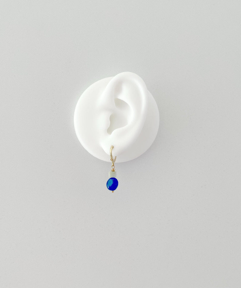 A single drop earring with a small gold huggie hoop, a pale green bead, and a deep blue oval jade gemstone, displayed on a white ear-shaped stand against a light gray background.