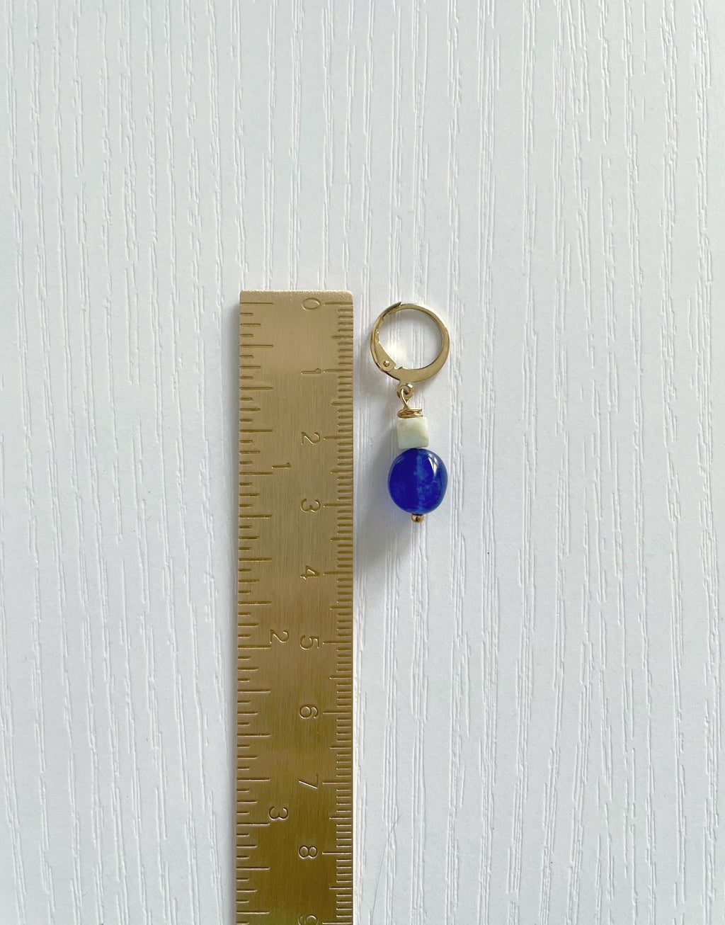 A small gold plated hoop earring with a single deep blue natural jade bead and a tiny sage green real amazonite stone, shown next to a metal ruler on a white textured background.
