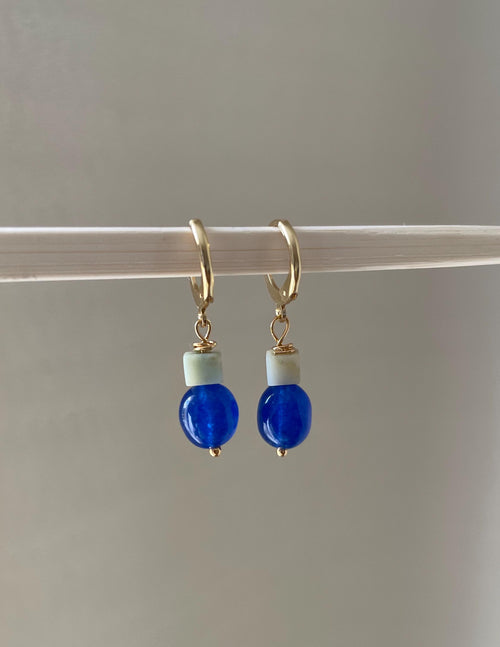 A pair of gold plated huggie hoop earrings with a small sage green amazonite bead and a glossy cobalt blue round bead dangling below, hanging from a white rod against a soft light gray background.