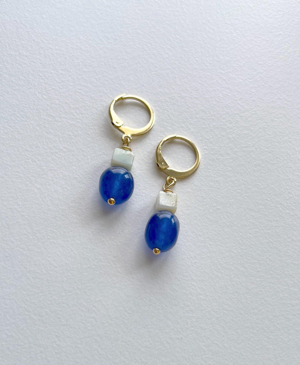 A pair of gold plated huggie hoop earrings with a small sage green square amazonite bead and a glossy royal blue oval natural jade bead dangling below, shown on a light gray textured background.
