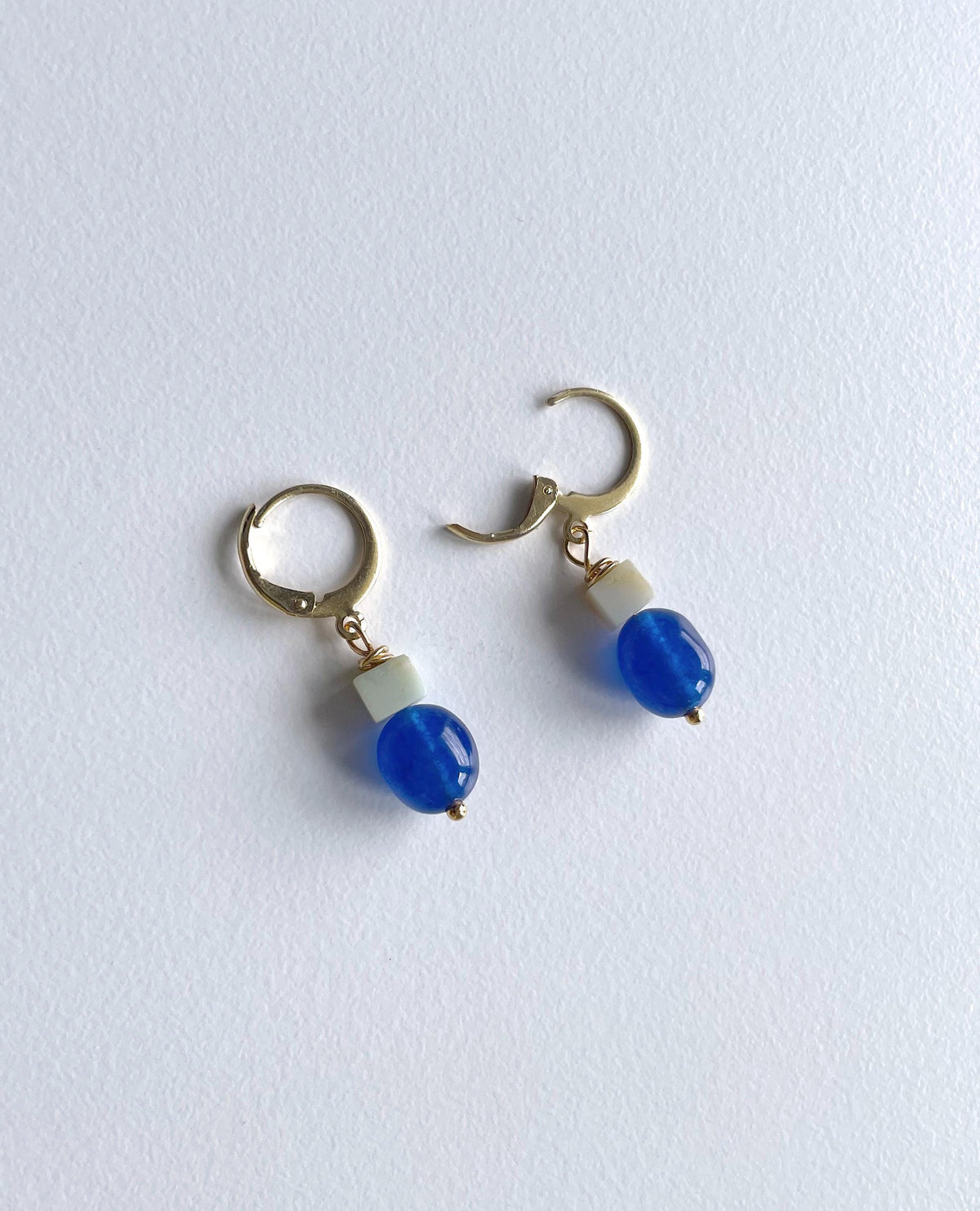 A pair of gold plated huggie hoop earrings with a small light green square bead and a dark blue jade bead dangling below, shown on a plain white background.