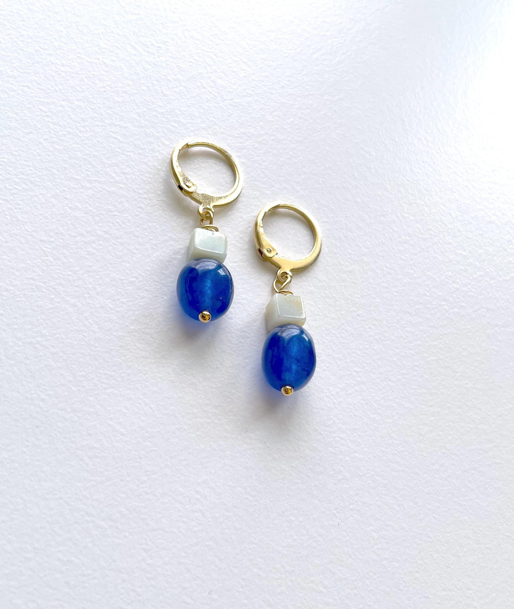 A pair of gold plated huggie hoop earrings with a dangling design featuring a small sage green square bead, a larger oval navy blue jade bead, and a tiny gold accent, displayed on a white textured background.