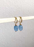 A pair of gold hoop earrings with oval light blue gemstone drops, hanging from a white rod against a textured off-white wall.