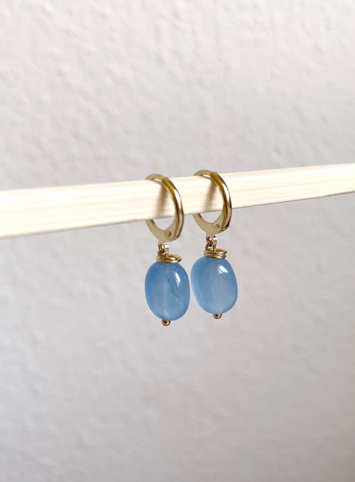 A pair of gold hoop earrings with oval light blue gemstone drops, hanging from a white rod against a textured off-white wall.