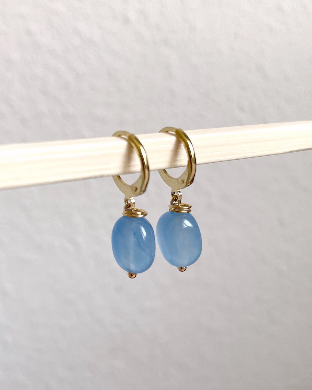 A pair of gold plated huggie hoop earrings with oval light blue natural gemstone drops, hanging from a white rod against a textured off-white wall.