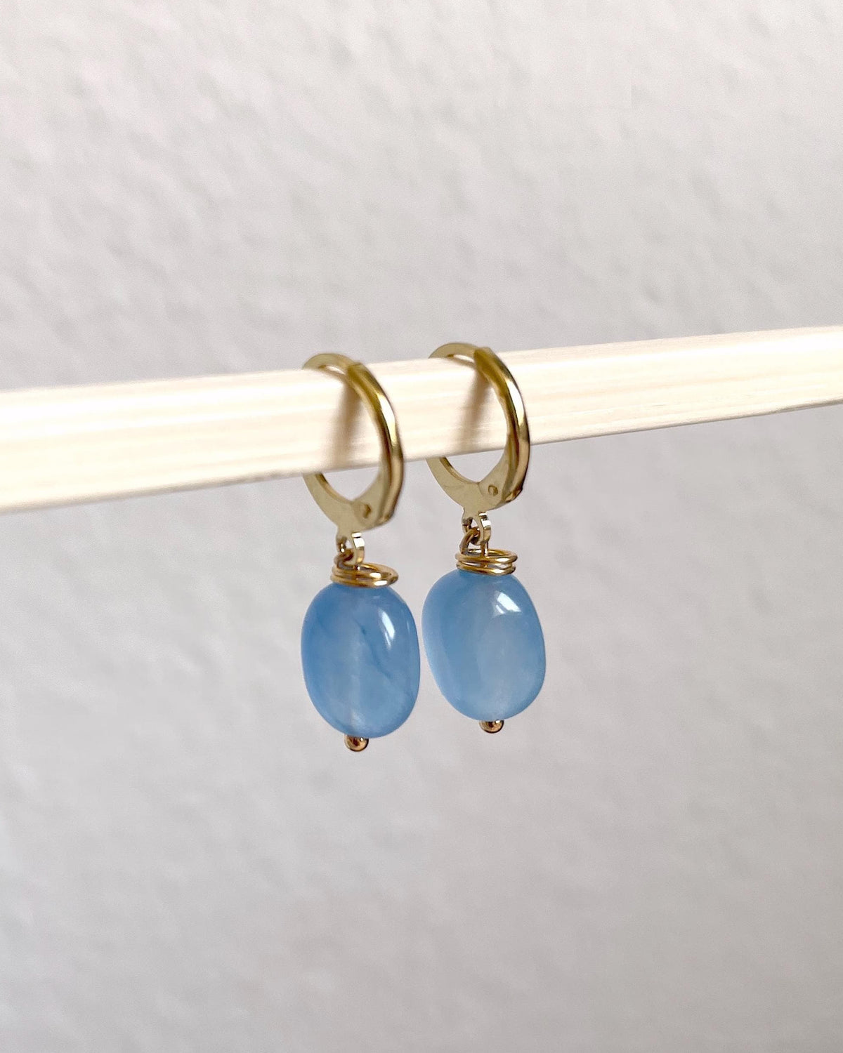 A pair of gold plated huggie hoop earrings with oval light blue natural gemstone drops, hanging from a white rod against a textured off-white wall.
