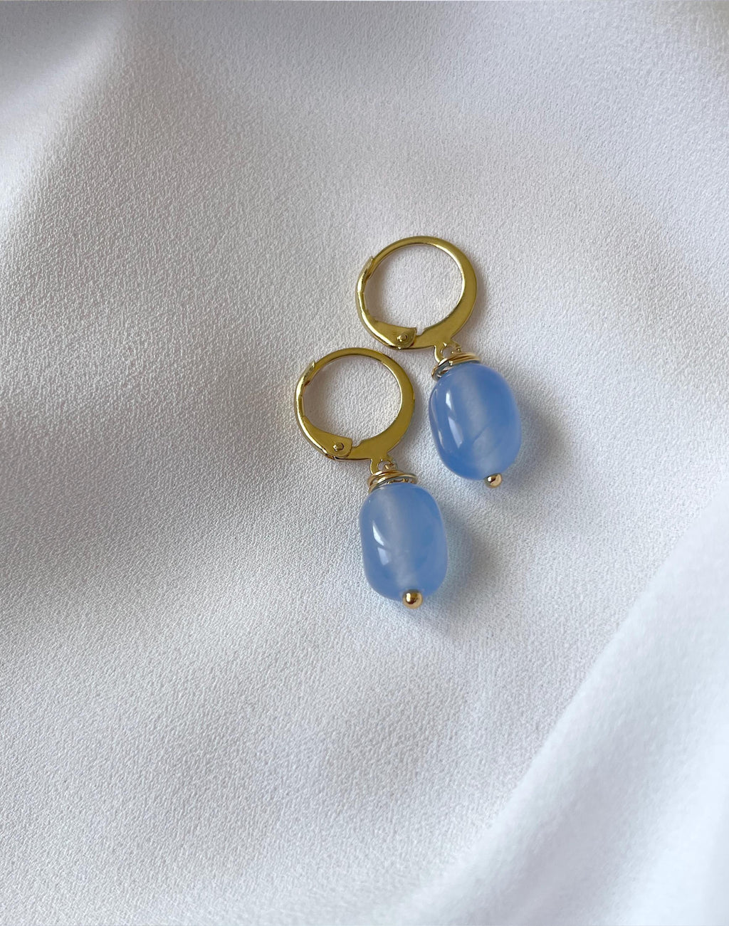 A pair of gold hoop earrings with dangling light blue oval beads, displayed on a soft white fabric background.