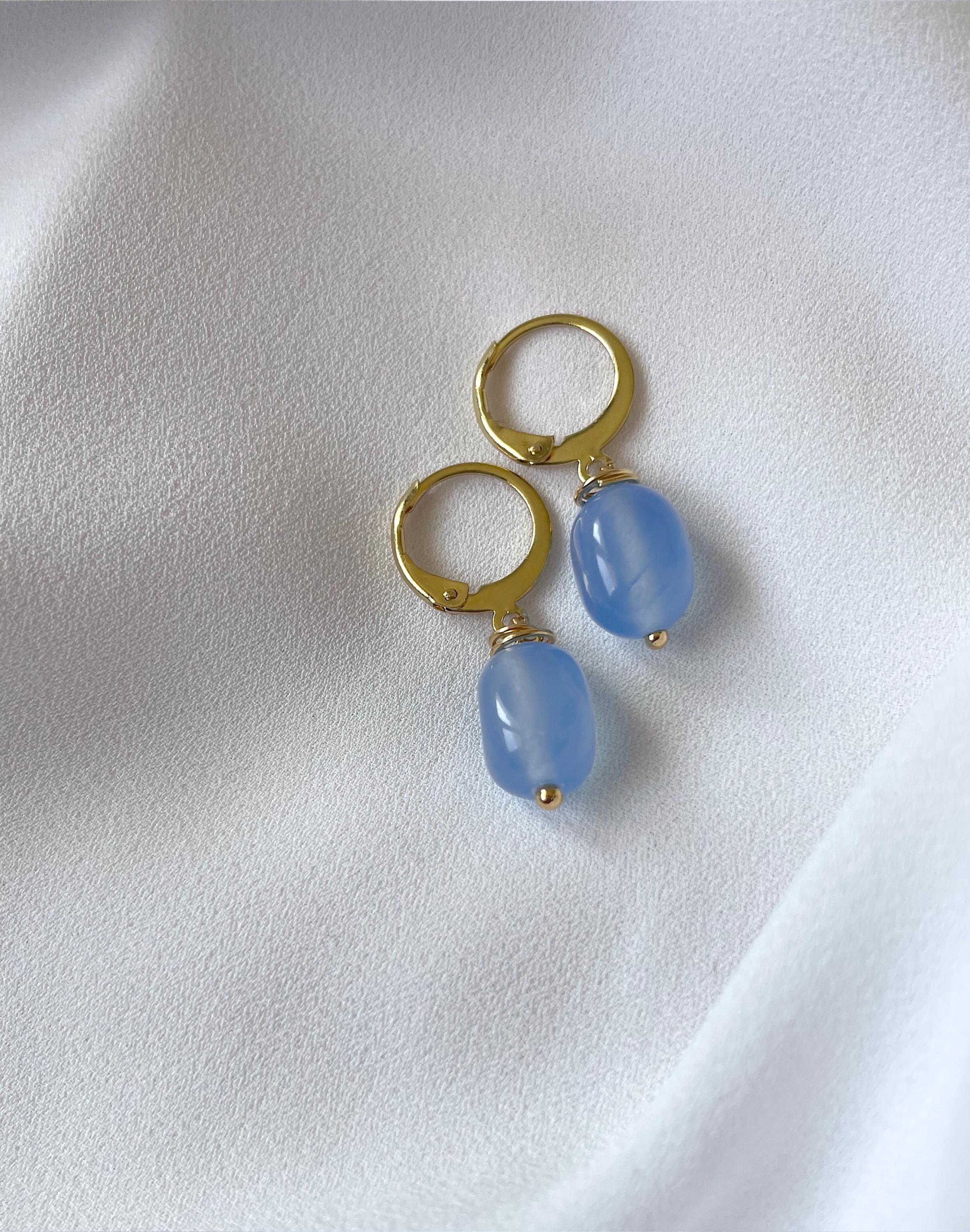 A pair of gold hoop earrings with dangling light blue oval beads, displayed on a soft white fabric background.