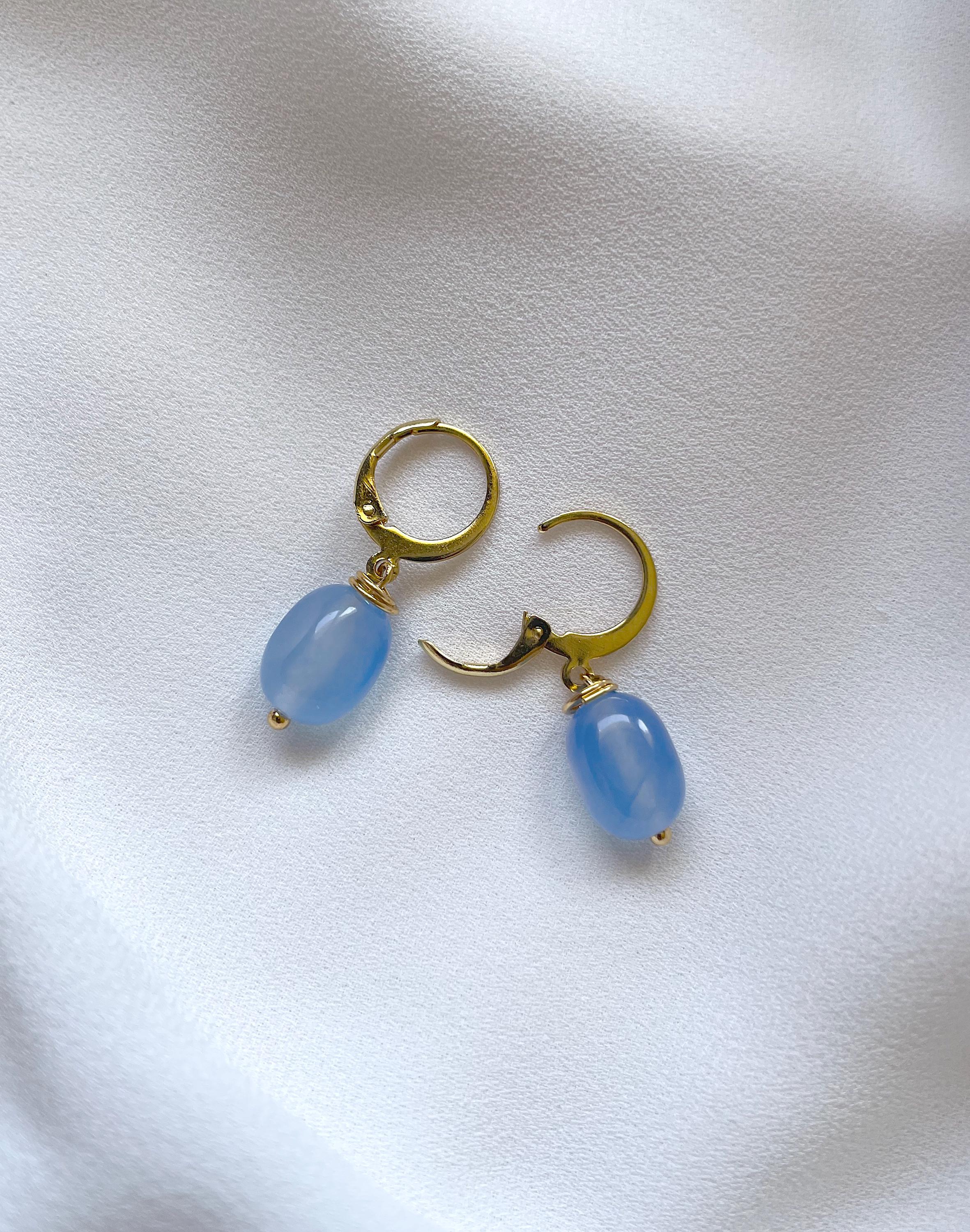 A pair of gold hoop earrings with smooth, light blue teardrop-shaped bead drops, displayed on a soft, textured white fabric background.