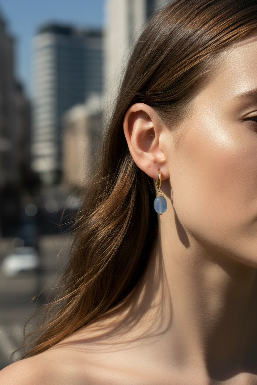 A close-up side profile of a woman wearing a delicate gold hoop earring with a small oval light blue stone dangle, set against a blurred city street background.